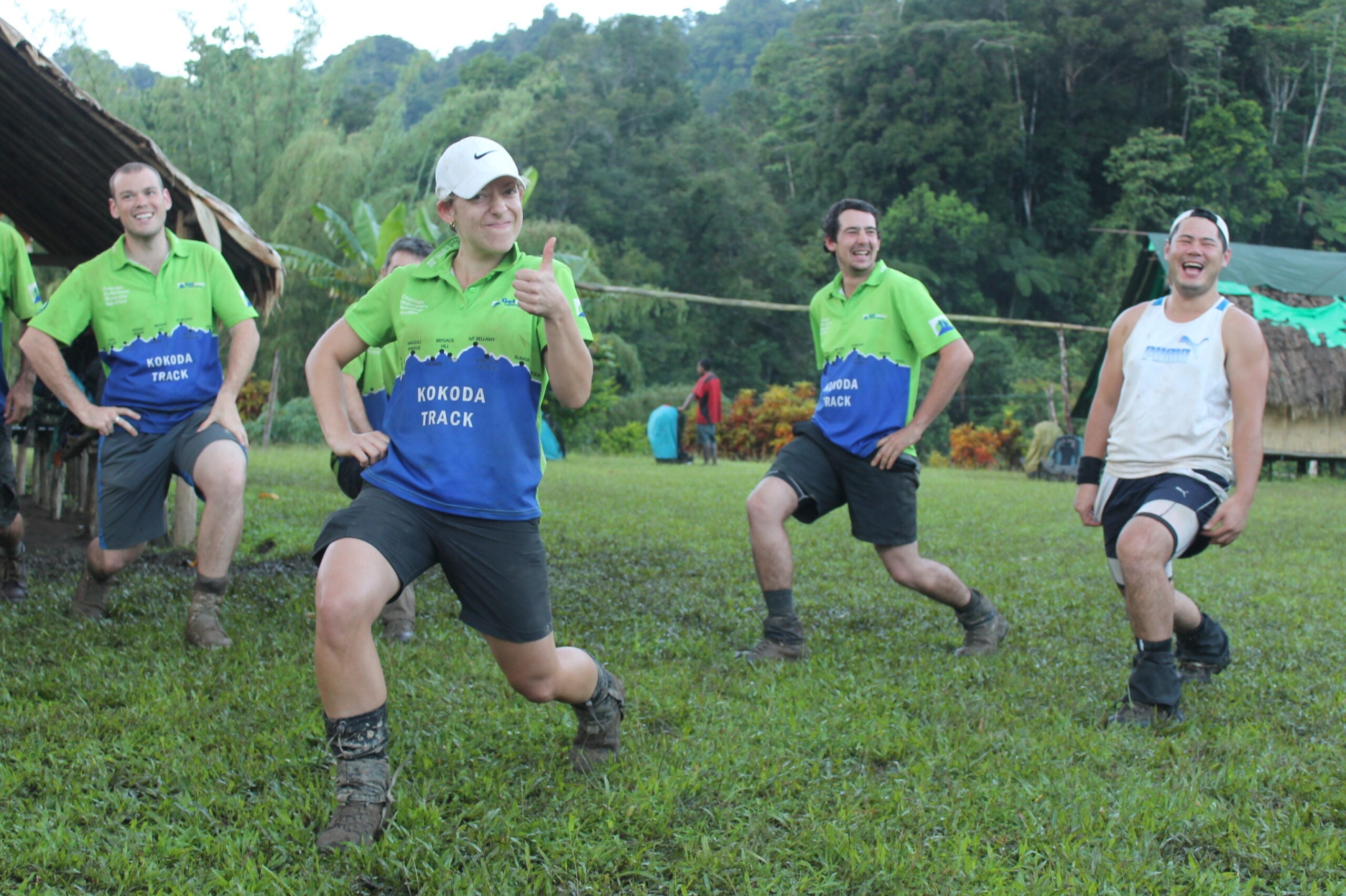 Four hikers are captured mid-lunge on a grassy field in a tropical setting. Three wear matching bright green and blue shirts labeled "Kokoda Track," while one woman in the front gives a thumbs-up to the camera. Lush green hills and a thatched structure are visible in the background.