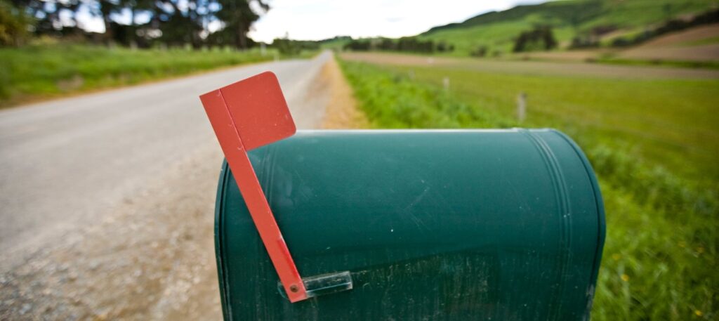 A green metal mailbox with a red flag up, situated on a quiet country road.