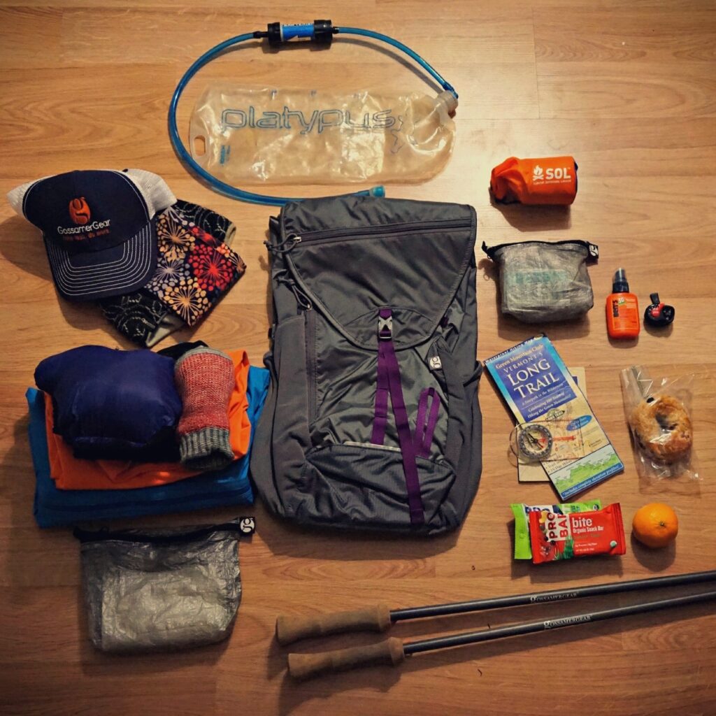 An overhead view of outdoor gear laid out on a wood surface, including a grey backpack with purple straps, trekking poles, a hydration bladder, a map of Vermont’s Long Trail, clothing, and snacks like an orange and granola bars.