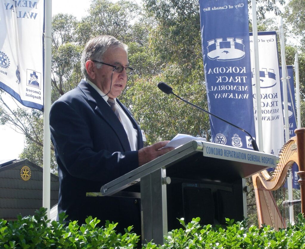 A side-profile shot of a man with grey hair and glasses speaking at an outdoor ceremony. He holds a piece of paper and stands behind a metal lectern. In the background, large blue and white banners for the "Kokoda Track Memorial Walkway" stand among green trees, and the corner of a wooden harp is visible to the right.