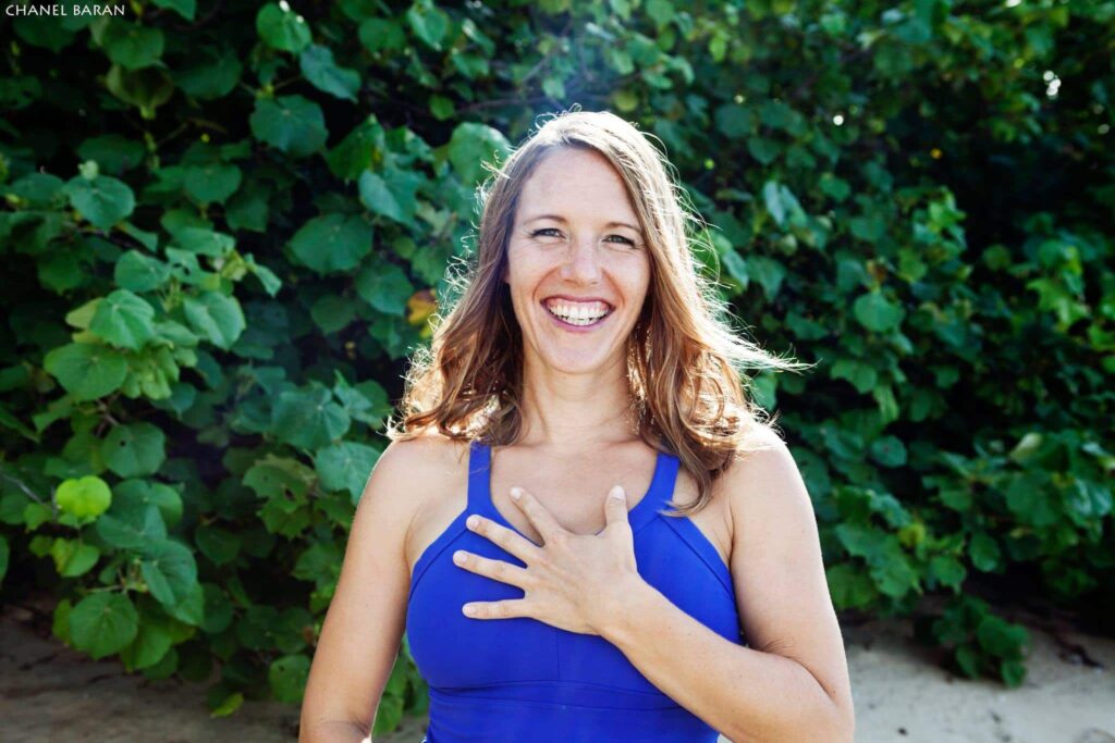 A bright outdoor portrait of a smiling woman with long brown hair, wearing a vibrant blue athletic top and resting her hand on her chest. She is posed against a lush background of green tropical leaves.