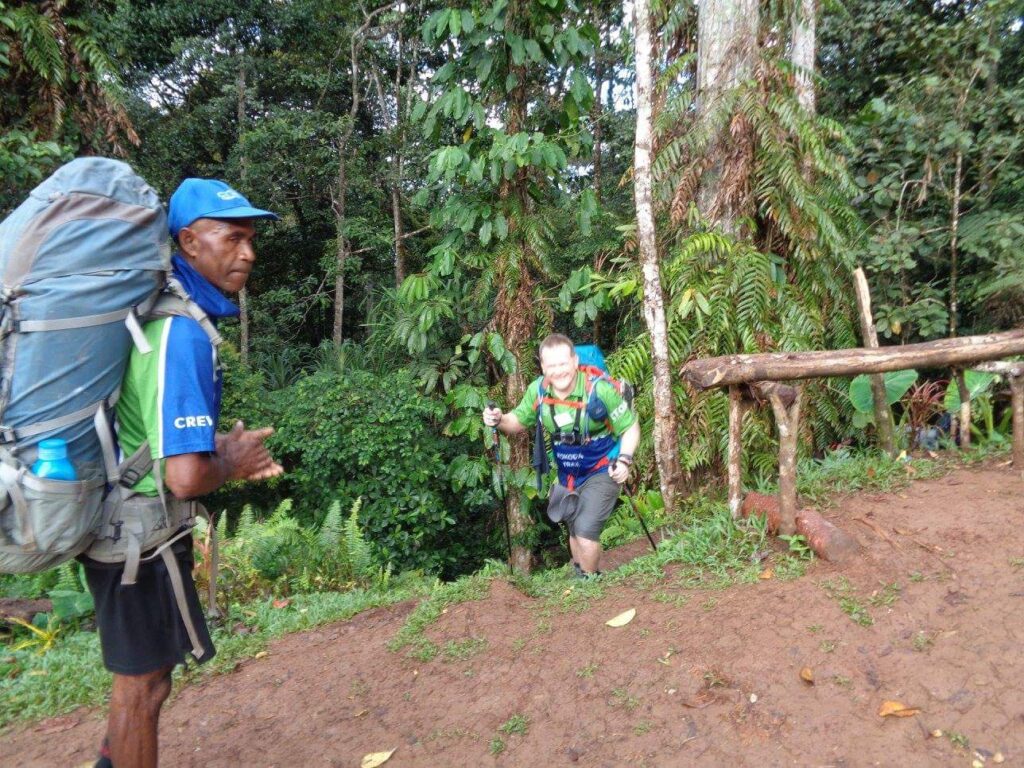 A local porter in a blue cap and green "CREW" shirt stands on a muddy slope, watching a trekker with trekking poles and a heavy blue backpack climb toward him through dense tropical forest.