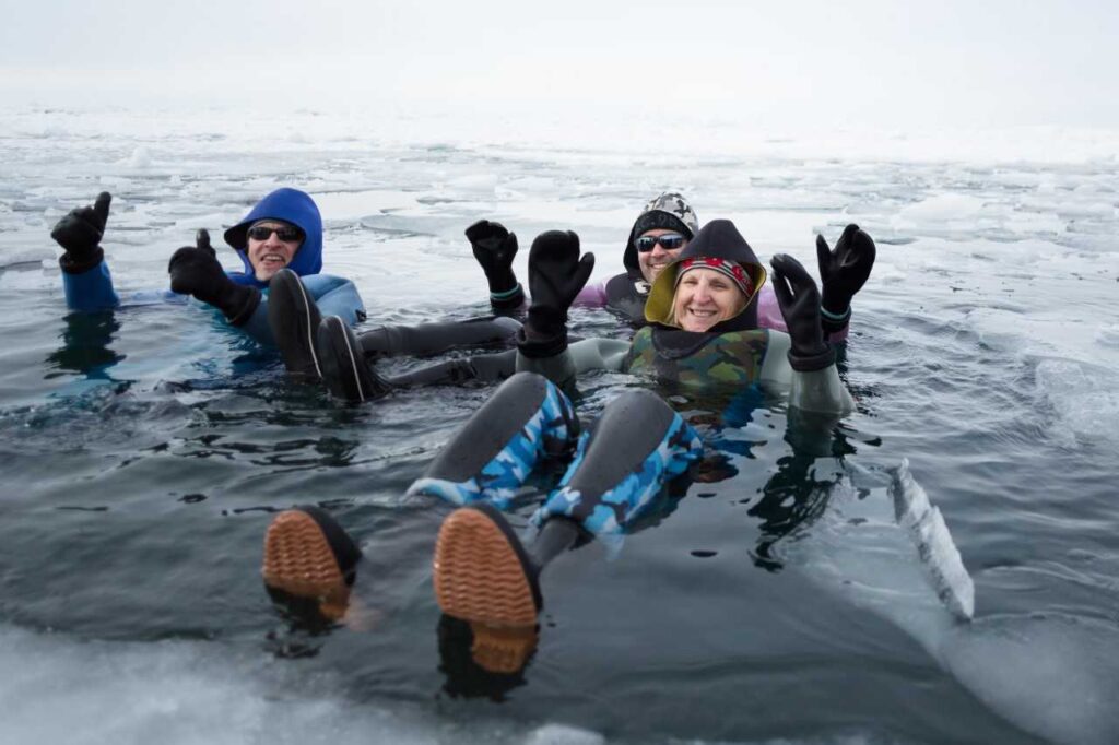 Four people wearing thick insulated dry suits float on their backs in dark, icy water surrounded by chunks of white ice.