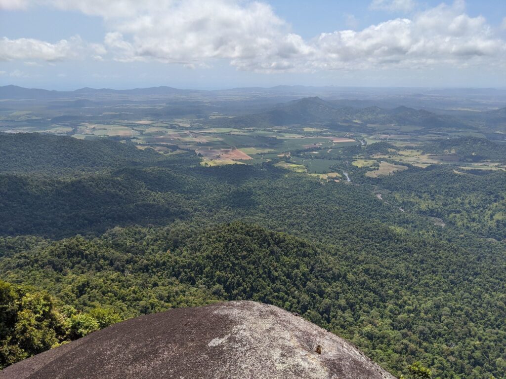 A high-angle view from a rounded, dark grey rocky peak looking out over an endless expanse of dense green tropical forest, with flat farmland and distant mountains visible on the horizon under a cloudy sky.