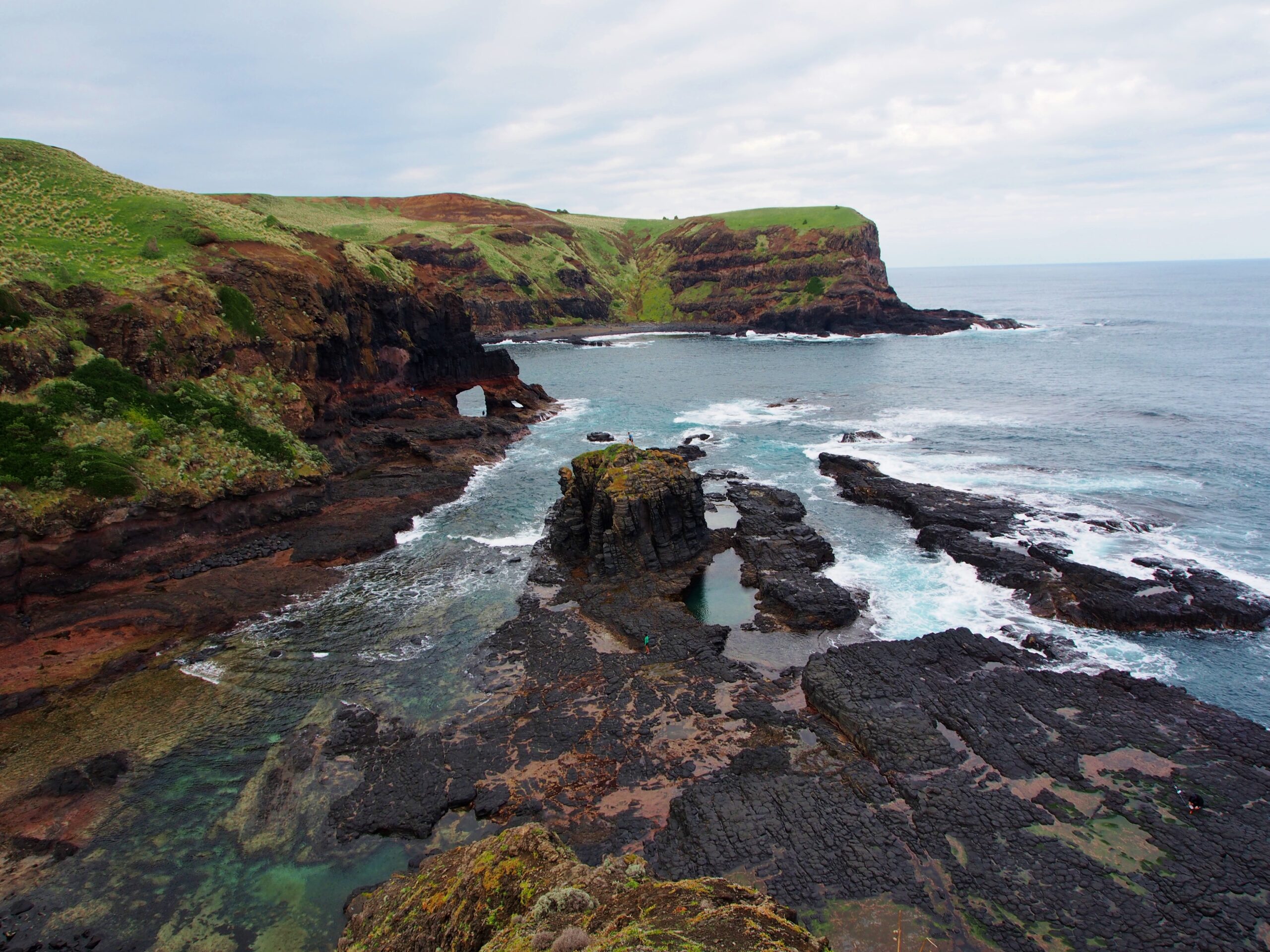 High-angle view of a rugged coastline featuring dark volcanic rock formations, tide pools, and a natural sea arch, with lush green hills meeting the crashing white waves of the ocean.