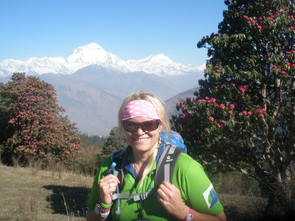A woman wearing a green trekking shirt, sunglasses, and a pink polka-dot headband smiles at the camera. She is surrounded by lush trees with vibrant pink flowers. In the background, sharp, white peaks rise above a blue mountain haze under a clear sky.