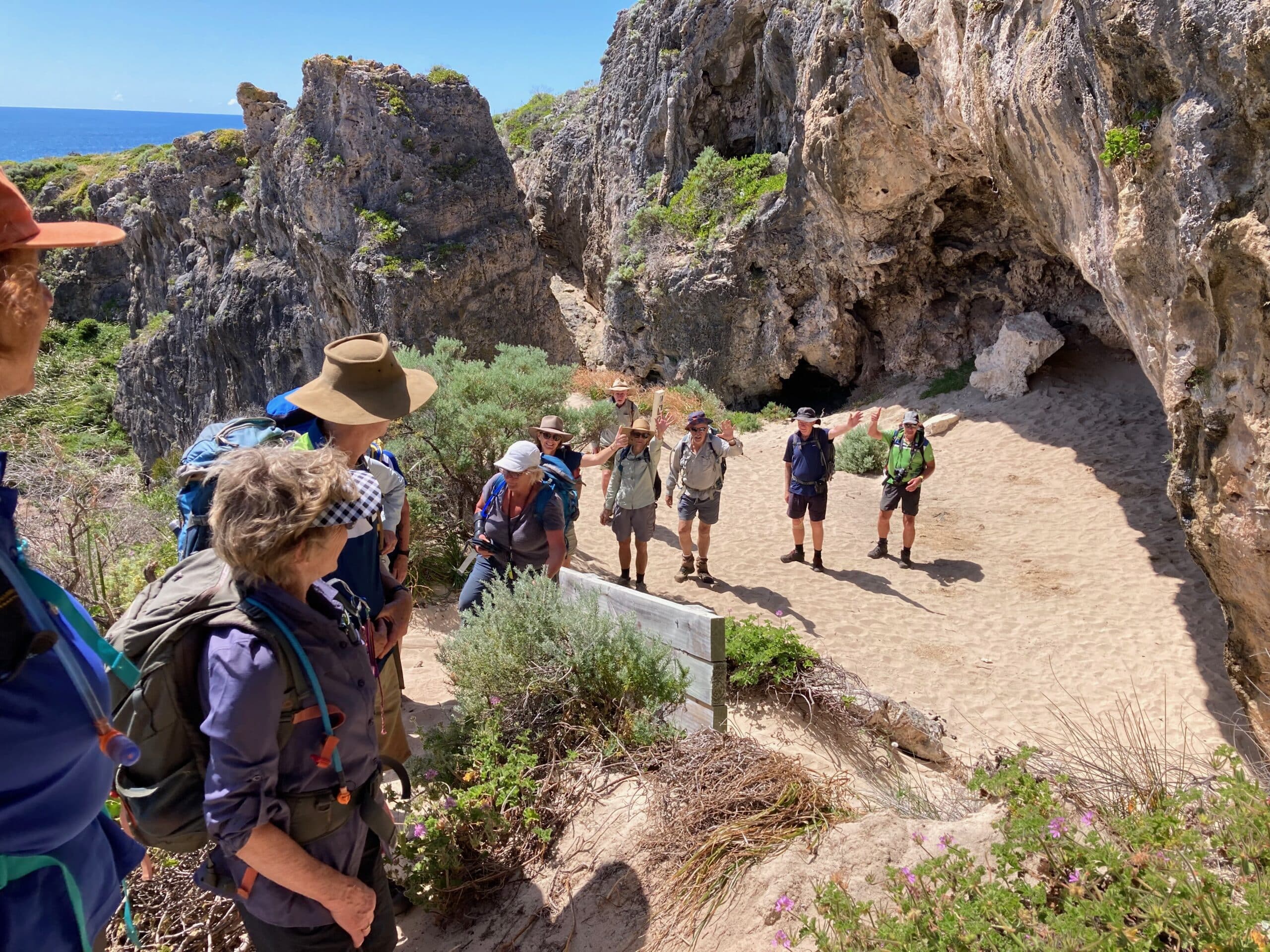 A group of hikers waving from a sandy area in front of a large, rugged sea cave along a rocky coastline.