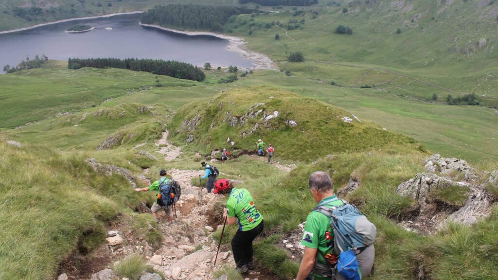 Hikers descending a rocky, grassy mountain path toward a large lake in a green valley.
