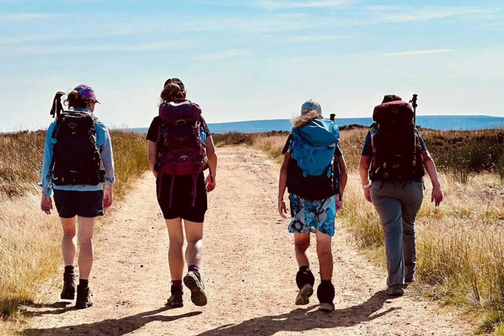 A view from behind of four hikers walking along a wide, dirt path through a grassy, open landscape. Each hiker carries a large backpack, and the horizon reveals a distant, flat-topped blue hill under a bright, clear sky.