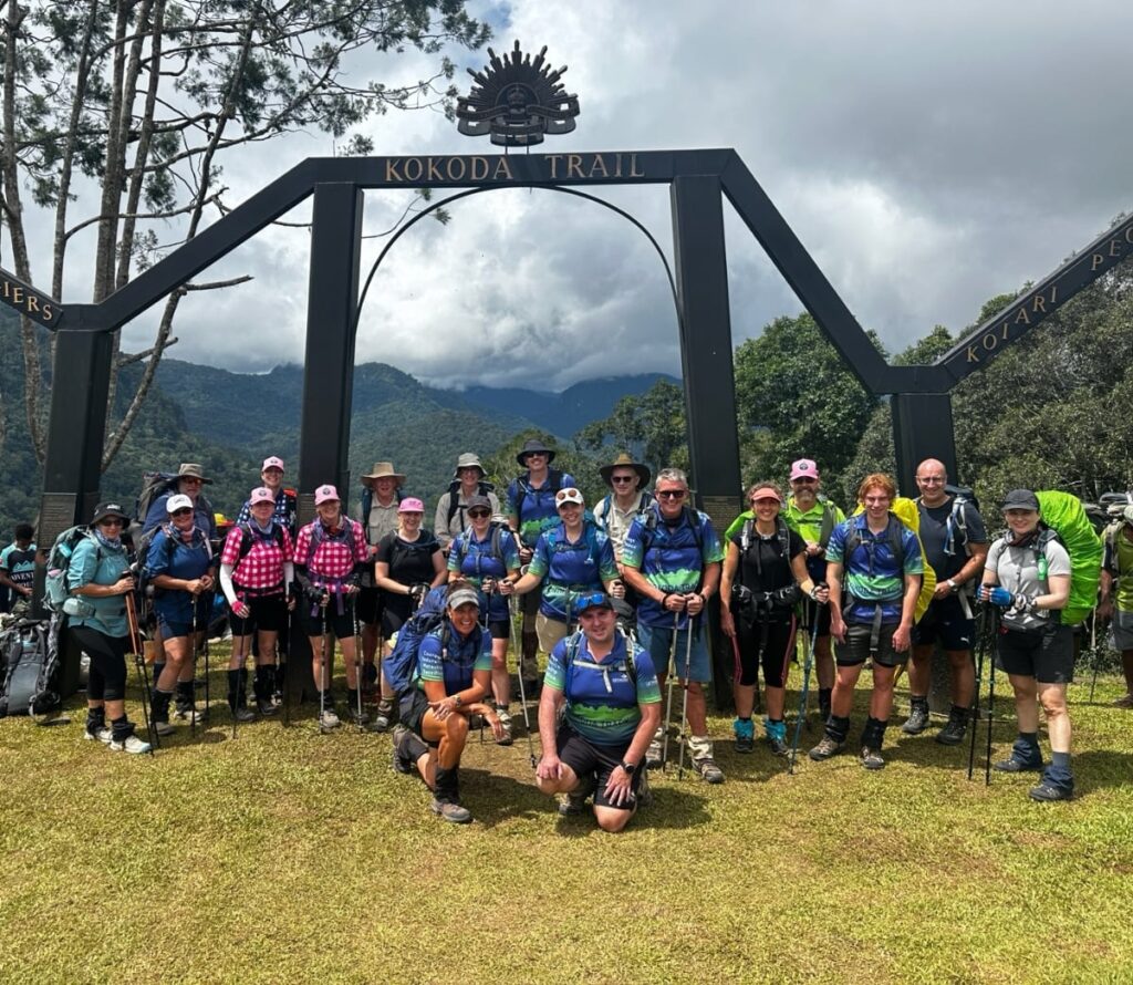 Group of hikers posing in front of large black archway labeled “KOKODA TRAIL”; side beams inscribed with “KOIARI” and “PERS”; hikers wear trekking gear including backpacks, hats, and walking poles; lush green mountains and cloudy sky form tropical backdrop; scene conveys camaraderie and adventure at historic trailhead.