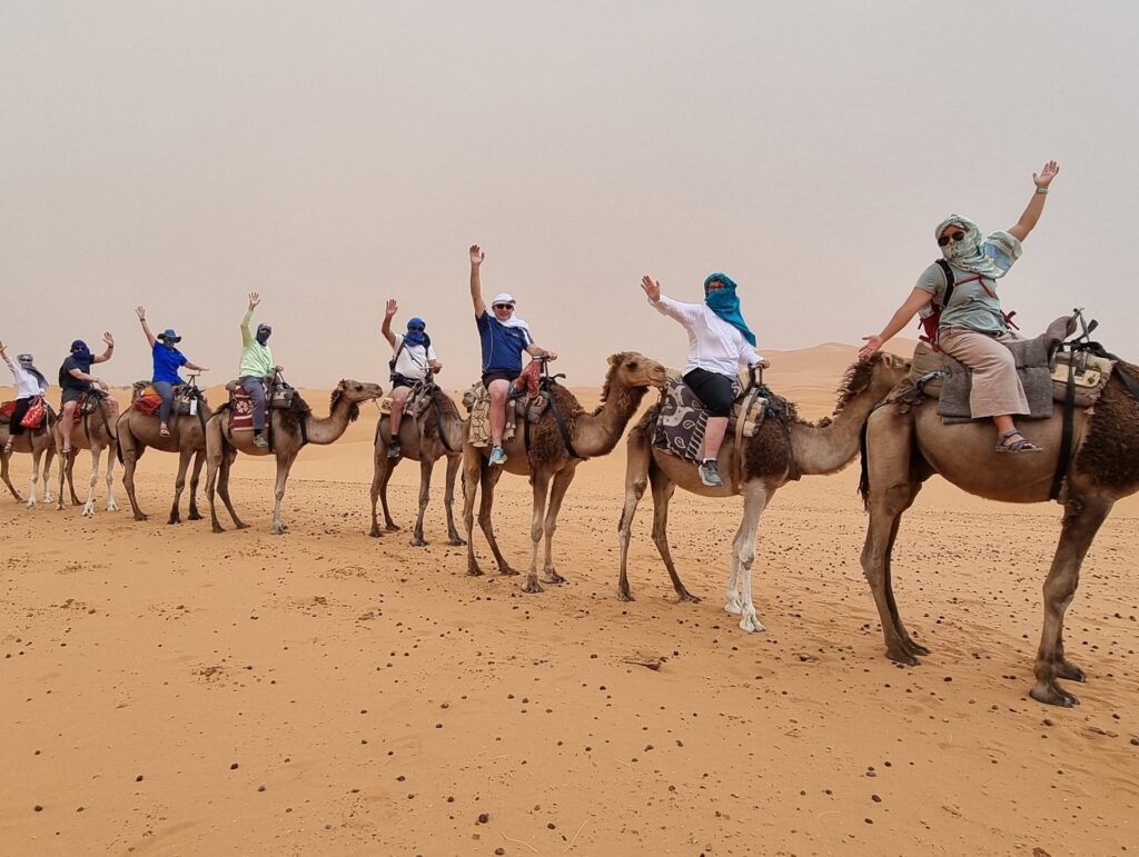 Group of eight people riding camels in desert; each person raises one arm, posing or waving; riders wear light protective clothing with headscarves and sunglasses; camels equipped with saddles and riding gear; vast sandy landscape with distant dunes under hazy sky.