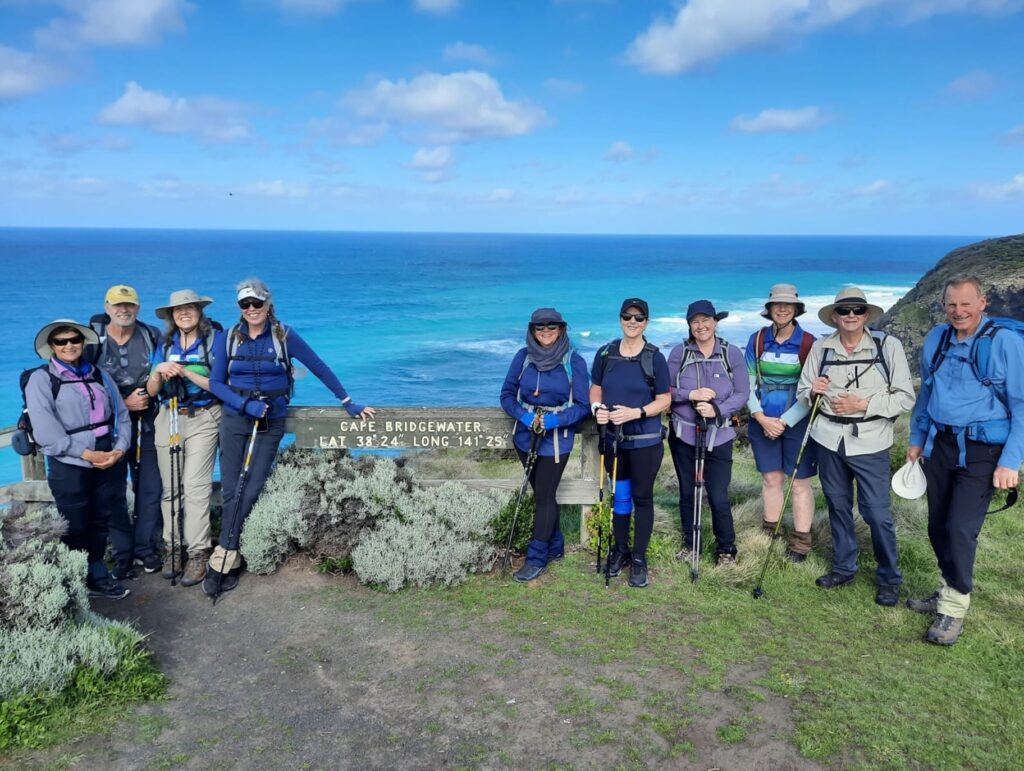 A group of nine hikers pose together on a grassy lookout with the ocean in the background. They wear outdoor gear including backpacks, hats, and walking sticks. Behind them stands a wooden sign reading "Cape Bridgewater LAT 38°24' LONG 141°25'," while waves crash against the rocky shore under a partly cloudy sky.