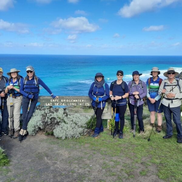 A group of nine hikers pose together on a grassy lookout with the ocean in the background. They wear outdoor gear including backpacks, hats, and walking sticks. Behind them stands a wooden sign reading "Cape Bridgewater LAT 38°24' LONG 141°25'," while waves crash against the rocky shore under a partly cloudy sky.