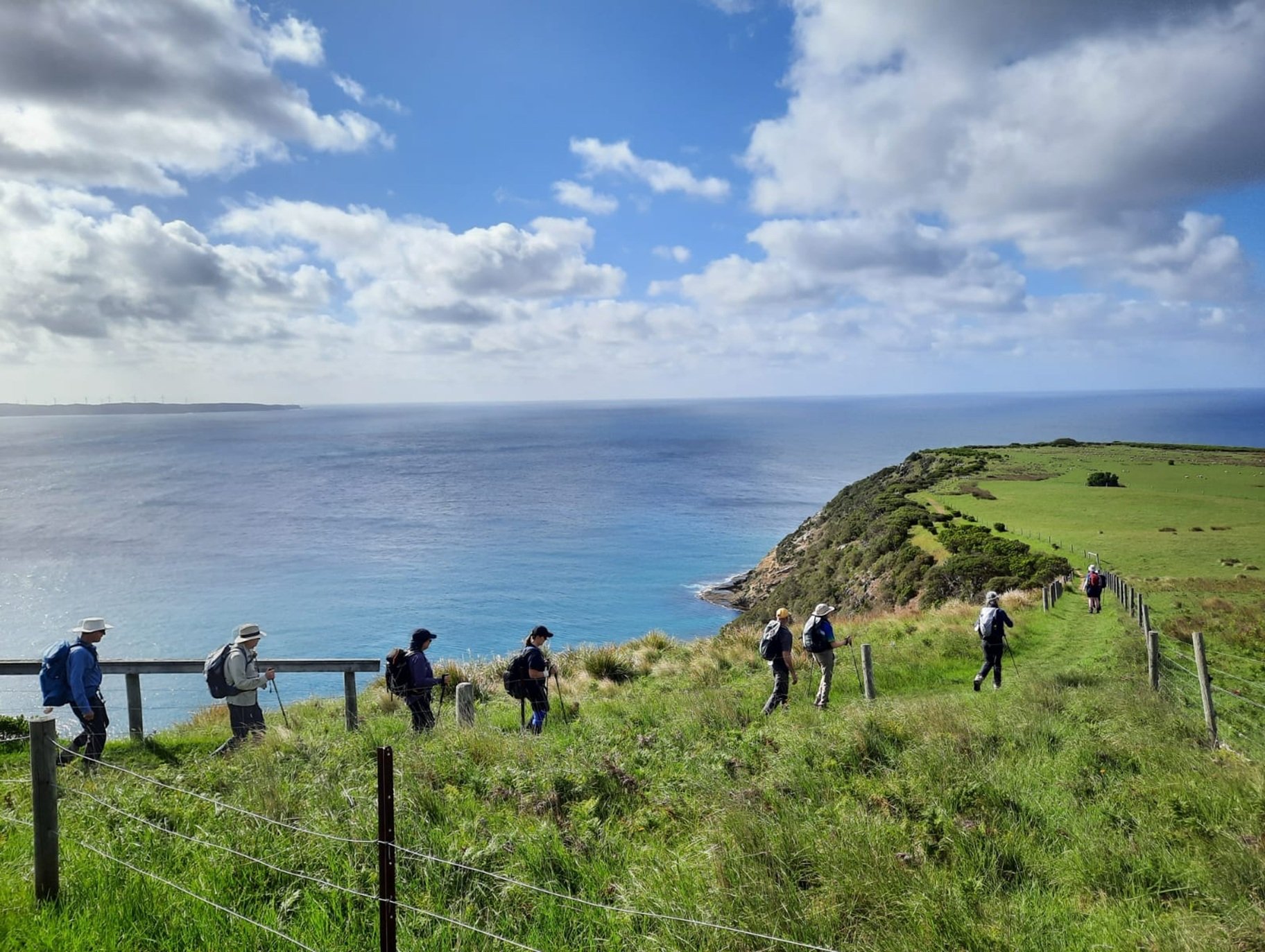 A group of hikers walk along a grassy cliffside trail overlooking a calm blue ocean. The scene is bathed in sunlight, with scattered clouds overhead. The hikers wear backpacks and sun hats, some using walking sticks, as they pass a wooden fence that borders the steep drop to the shore below. The terrain combines lush greenery and rocky edges, creating a scenic coastal landscape.