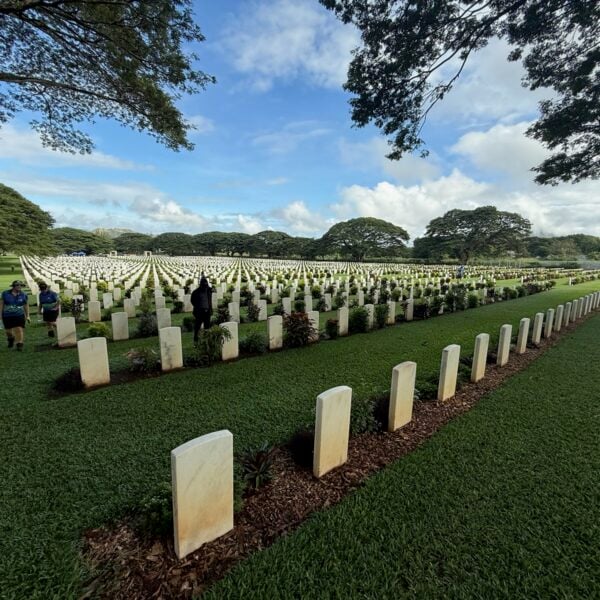 The image shows a large cemetery with neatly arranged gravestones in Kokoda. The graves are aligned in rows across a well-maintained grassy field. There are a few people walking between the rows of gravestones, and tall trees line the background under a partly cloudy sky. The environment appears peaceful and serene.
