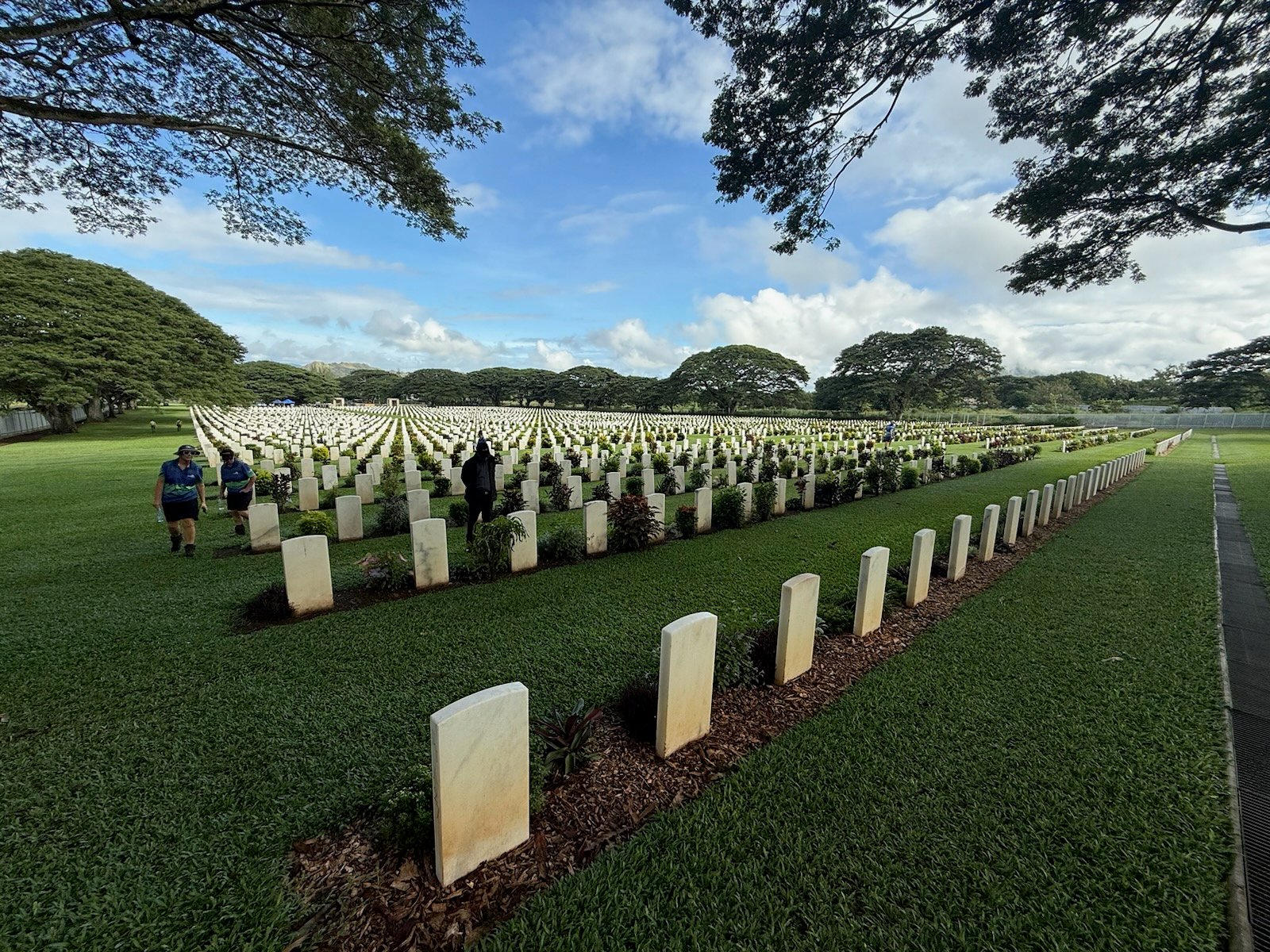 The image shows a large cemetery with neatly arranged gravestones in Kokoda. The graves are aligned in rows across a well-maintained grassy field. There are a few people walking between the rows of gravestones, and tall trees line the background under a partly cloudy sky. The environment appears peaceful and serene.