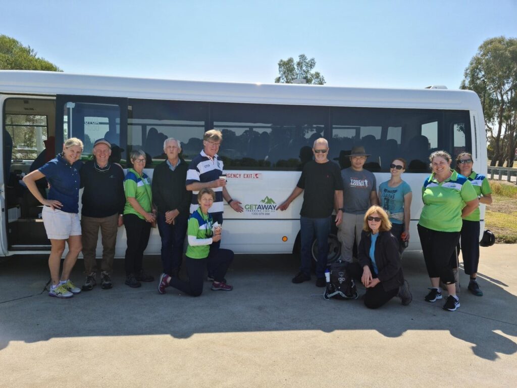 Group of thirteen people posing in front of a white passenger bus with tinted windows; bus displays “GETAWAY” and phone number “1300 576 683,” with “EMERGENCY EXIT” above; individuals dressed casually, some in matching green and blue shirts, outdoors on a sunny day with trees and clear sky in the background.