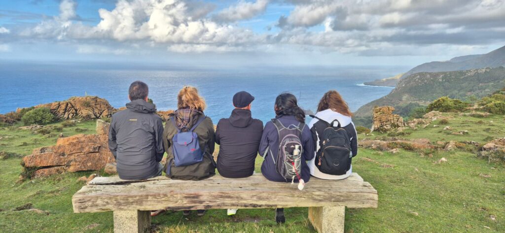 Five hikers with backpacks sitting on a rustic wooden bench, facing the ocean and rugged cliffs under a partly cloudy sky; grassy and rocky terrain in the foreground, with distant hills along the coastline.