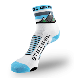 A white Steigen athletic sock featuring a detailed landscape graphic on the cuff depicting grey mountains with snow caps and small green evergreen trees. The toe and heel are light blue, and the brand name is printed vertically in black on the foot next to light blue and black arch stripes.