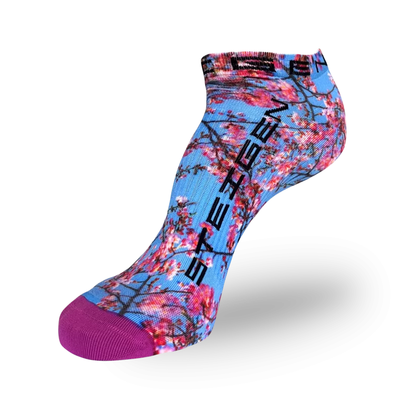 A low-cut athletic sock featuring a light blue base covered in a pink cherry blossom print with dark brown branches. The brand name "STEIGEN" is printed in bold black text vertically down the side. The sock has a solid purple reinforced toe.