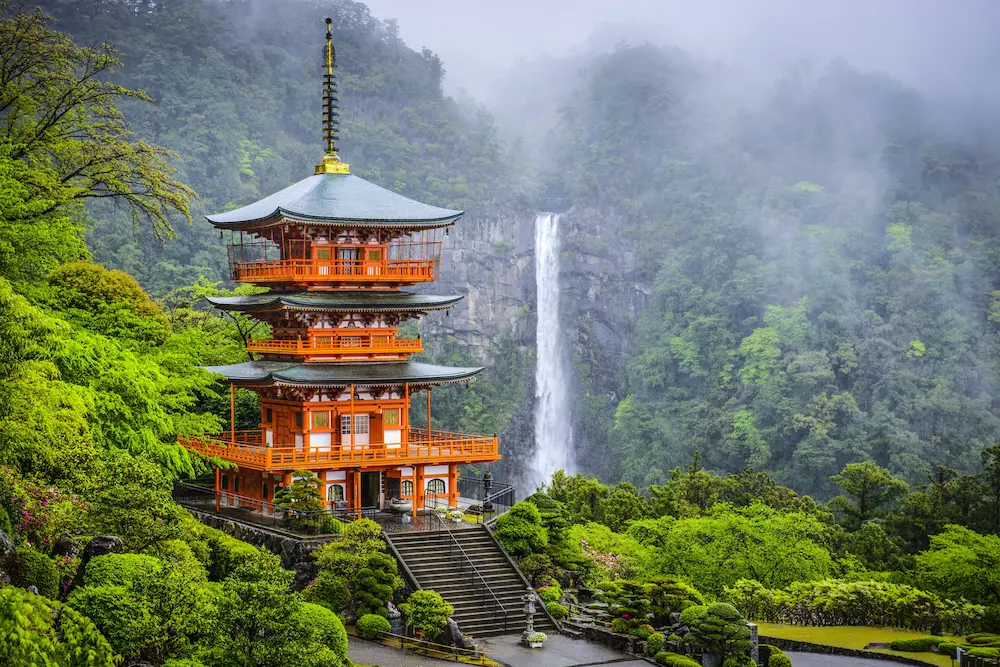 Traditional Japanese pagoda‑style temple painted orange and white, with multiple tiers and curved roofs, set among manicured gardens and stone steps; lush greenery and mist‑covered mountains in the background, with a tall waterfall cascading down a rocky cliff.