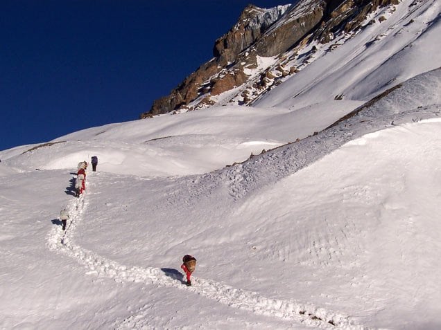 Group of trekkers walking in single file along a snow-carved path in rugged high-altitude mountains, carrying loads on their backs, with steep rocky slopes and clear deep blue sky in the background.