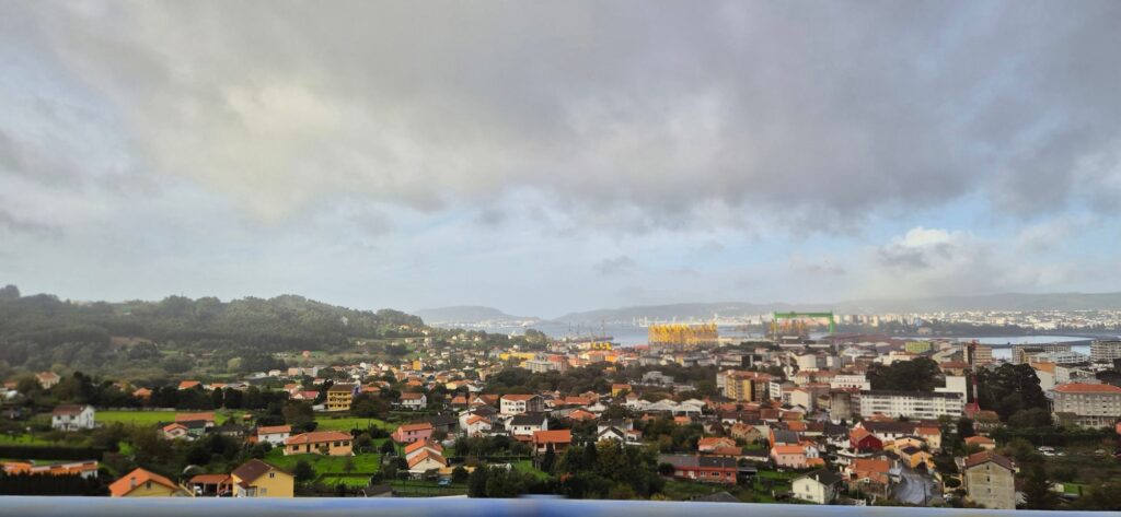 Panoramic view of a town with red and orange rooftops in the foreground, dense buildings and port cranes in the middle ground, and forested hills with urban development across a bay under a partly cloudy sky.