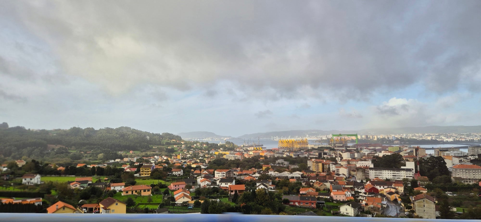 Panoramic view of a town with red and orange rooftops in the foreground, dense buildings and port cranes in the middle ground, and forested hills with urban development across a bay under a partly cloudy sky.