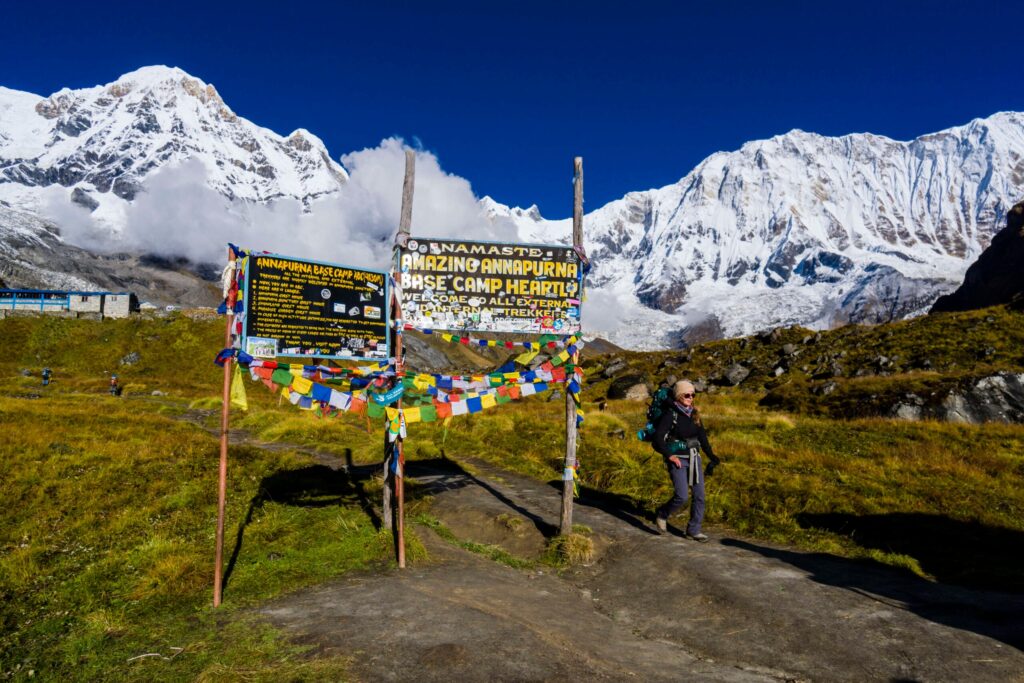 A hiker walking past a colorful "Welcome to Annapurna Base Camp" sign with massive snow-capped Himalayan mountains in the background under a deep blue sky.