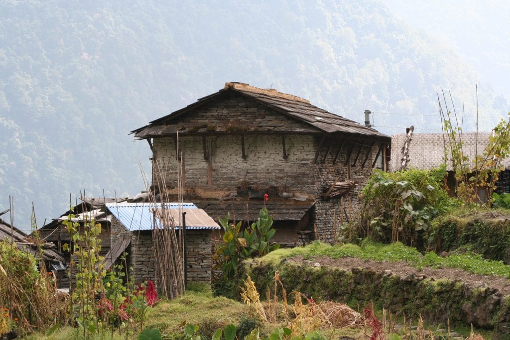 Traditional stone house with stacked stone walls and wooden roof beams in a lush mountainous rural area, surrounded by smaller structures and greenery, with misty forested slopes in the background.