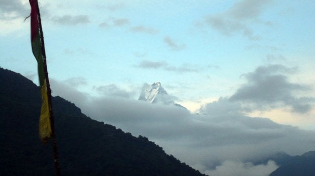 A sharp, triangular snow-capped mountain peak peeking through a thick layer of white clouds at dawn, with a dark silhouetted hillside and prayer flag in the foreground.