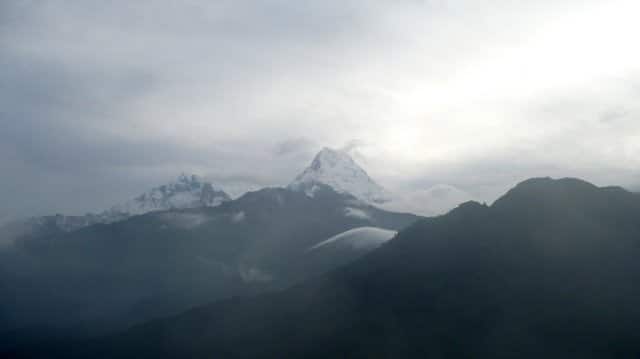 A moody, high-altitude landscape featuring a large, pyramid-shaped snowy peak in the center and a secondary jagged peak to its left. The mountains are partially shrouded in swirling white clouds and heavy grey mist. In the foreground, the dark, shadowed ridges of lower mountains create a stark contrast against the hazy, atmospheric sky.