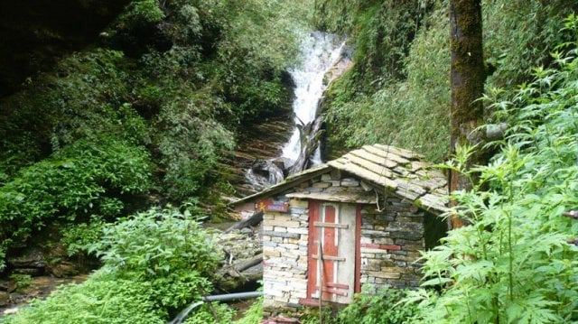 Small stone hut with a wooden door and slanted stone-tiled roof, set in a lush forest with a waterfall cascading down a rocky slope behind it, surrounded by dense vegetation.