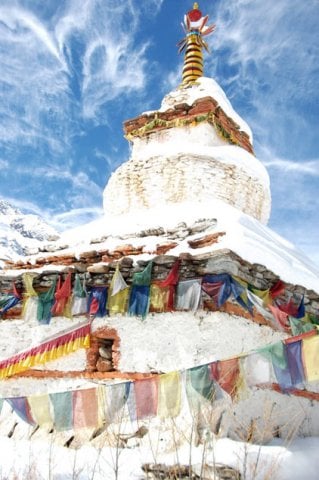 A white, traditional Buddhist chorten covered in fresh snow and draped with colorful prayer flags under a bright blue, wispy sky.