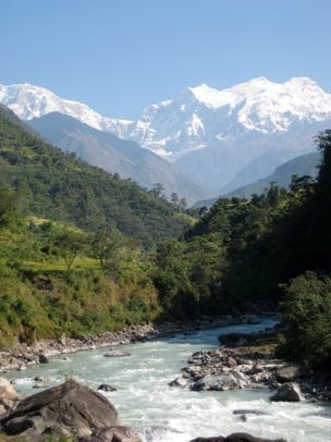 Scenic landscape with a flowing river in the foreground, surrounded by lush green hills and forest, with towering snow-capped mountains rising in the background under a clear blue sky.