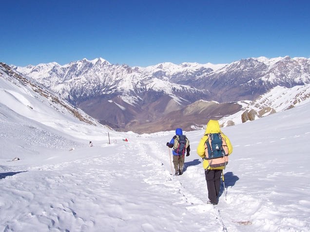 Two hikers in bright jackets trek down a steep, snow-covered mountain pass toward a vast range of jagged, snow-capped peaks.