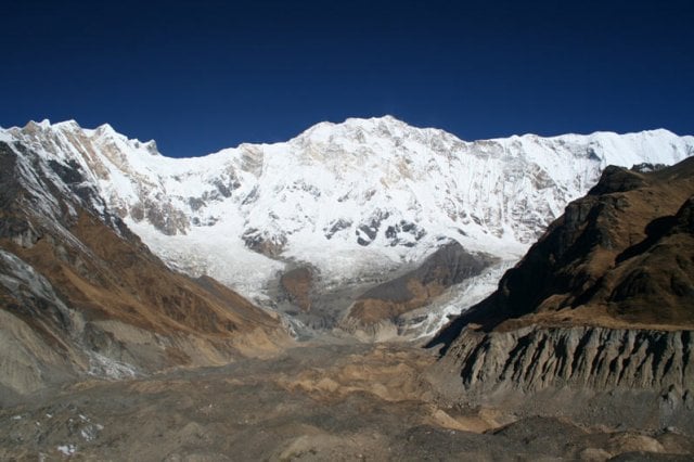 A wide-angle view of a rugged glacial valley with rocky moraine in the foreground, leading up to a massive, snow-covered mountain wall under a dark blue sky.