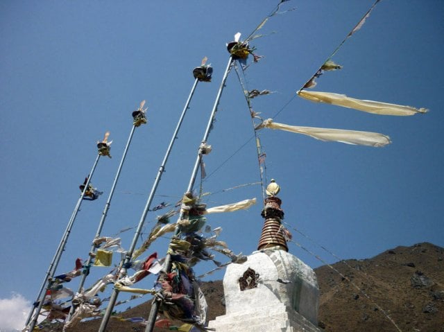 Traditional Tibetan stupa with white dome and golden spire, surrounded by tall poles draped in colorful prayer flags fluttering in the wind, set against a clear blue sky and mountainous backdrop.