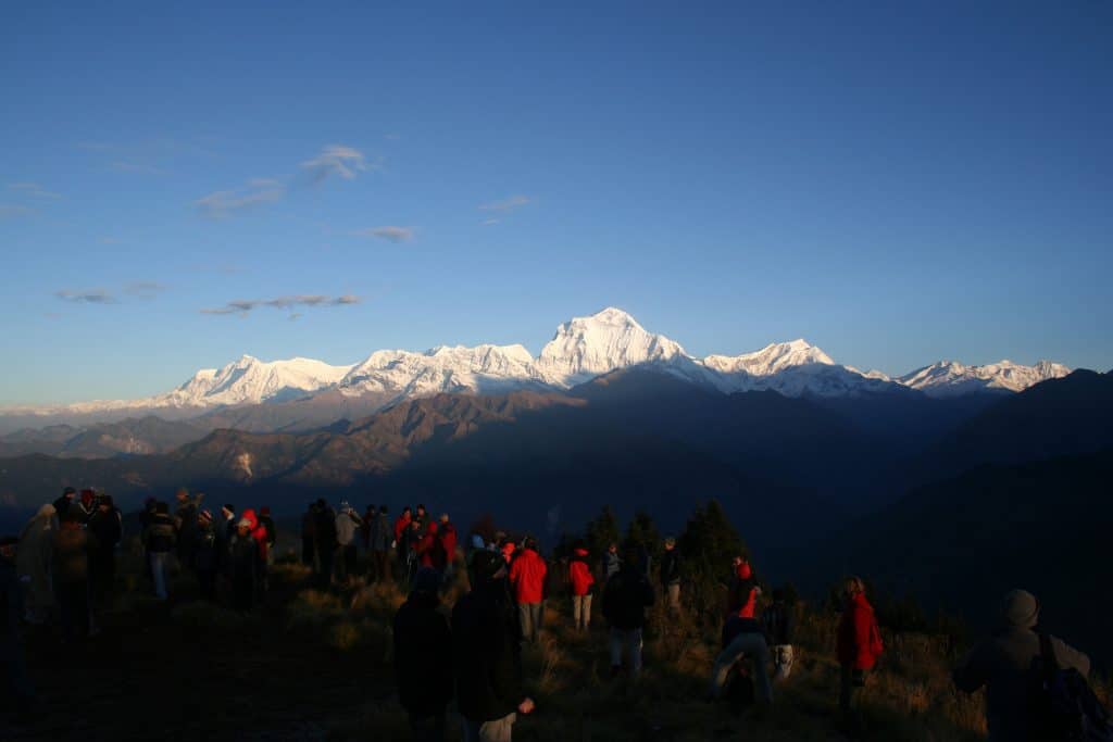 A wide panoramic view of snow-capped Himalayan peaks under a clear blue sky, with a group of silhouetted trekkers in the foreground.