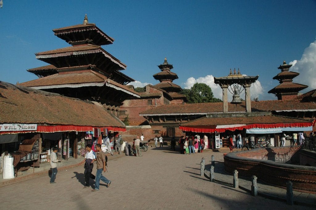 A wide view of a sunlit plaza in Patan, Nepal. The scene is dominated by several traditional Newar temples with tiered brown roofs and ornate wooden carvings. To the right, a large ceremonial bell hangs from a stone structure. People walk through the open square, which is lined with low-slung shops, including one with a sign for a tailor. The sky is a deep blue with soft white clouds in the distance.