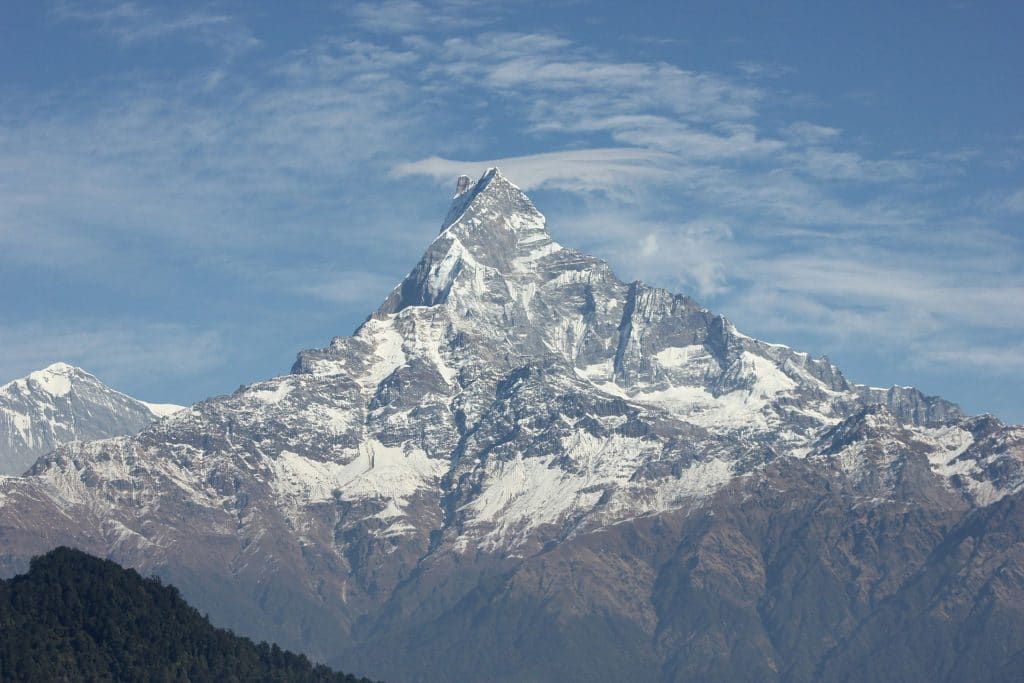Majestic snow-covered mountain peak with sharp summit and rugged slopes, rising above dense green forested hills under a clear blue sky with scattered clouds.