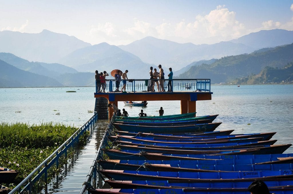 A row of colorful blue wooden boats docked at a lakeside pier with people standing on an elevated platform against a backdrop of hazy mountains.