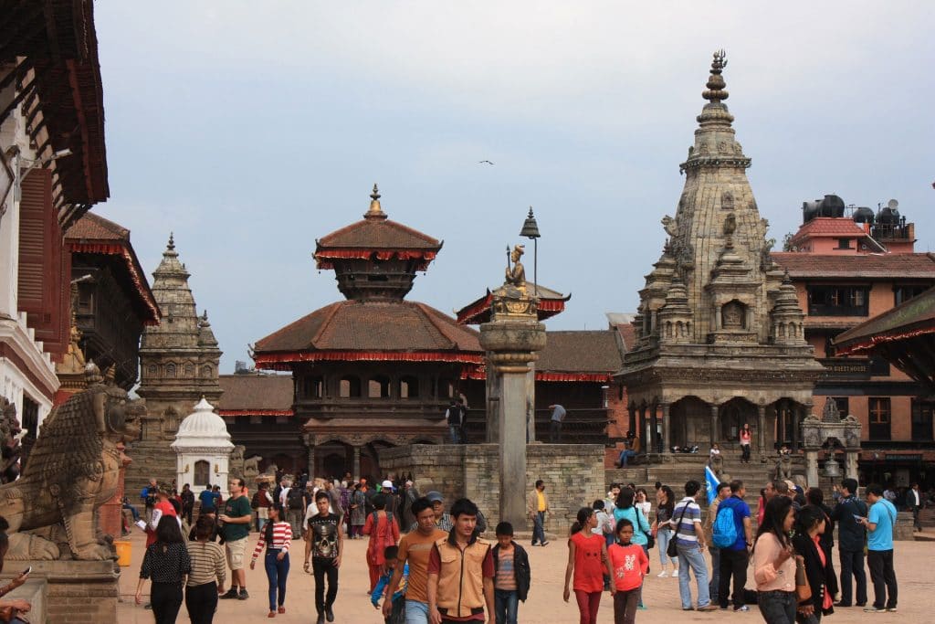 Bustling Patan Durbar Square in Lalitpur, Nepal, with pagoda-style temples, ornate woodwork, stone pillar topped with a statue, and crowds of locals and tourists among historic sculptures and architecture.