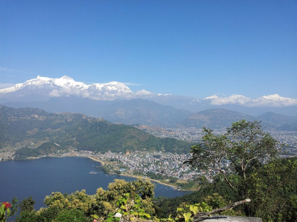 A high-angle panoramic view of a large blue lake bordering a densely populated city, with lush green hills in the foreground and massive snow-capped mountains in the distance.