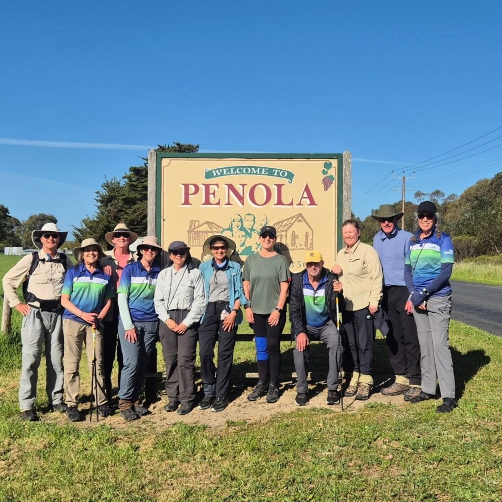 Group of twelve people in hiking gear standing in front of a large roadside sign reading “WELCOME TO PENOLA”; illustrated sign includes people, buildings, and a bunch of grapes symbolizing vineyards; rural setting with grass, trees, and power lines under a clear blue sky.