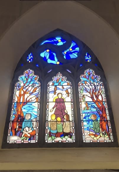 Stained glass window divided into three vertical panels; central panel shows robed figure standing with three seated children, suggesting teaching or storytelling; left and right panels depict children under trees with colorful leaves, birds, and a flowing river; upper section features abstract blue and white swirling patterns with yellow accents, resembling sky or heavens.