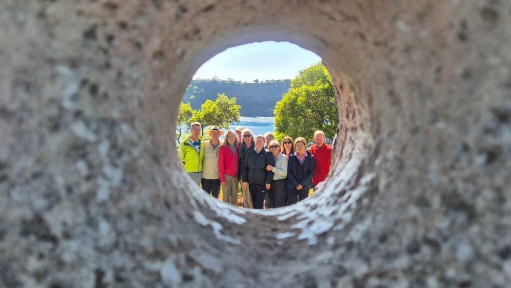 Group of people posing outdoors, framed through a circular hole in a textured stone or concrete surface; background shows green trees, a body of water, and a forested hill under a clear blue sky.