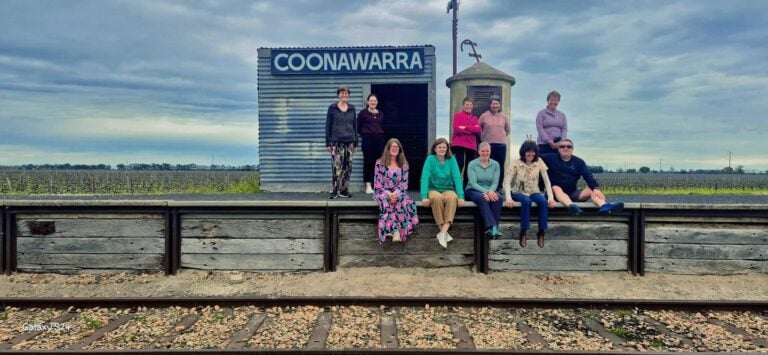 Group of ten people posing on a rustic train station platform labeled “COONAWARRA”; corrugated metal building and cylindrical structure beside the platform; wooden planks elevate the platform next to railway tracks; background shows expansive vineyard under a cloudy sky; group casually dressed, some standing and others sitting on the edge of the platform.