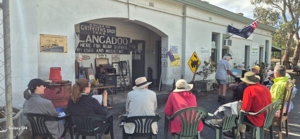 Group of people seated outdoors in front of a historic roadside building decorated with vintage signs and memorabilia; Australian flag displayed, with a man standing and giving a presentation to the audience in plastic chairs; setting suggests a community gathering or educational event at a heritage site.
