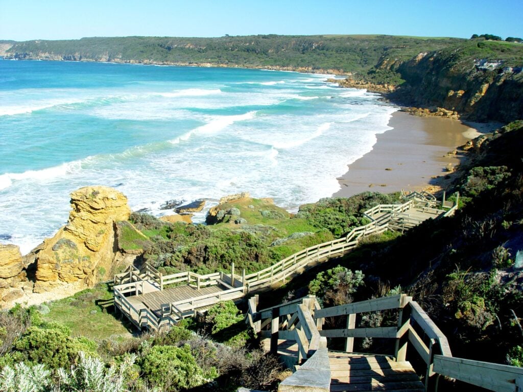 A high-angle view of a rugged coastline. A light-colored wooden boardwalk with several zig-zagging staircases and square observation decks descends a steep, scrub-covered cliff. To the left, a golden rock formation stands near the white surf of turquoise ocean waves. A long, sandy beach stretches toward distant green headlands under a clear sky.